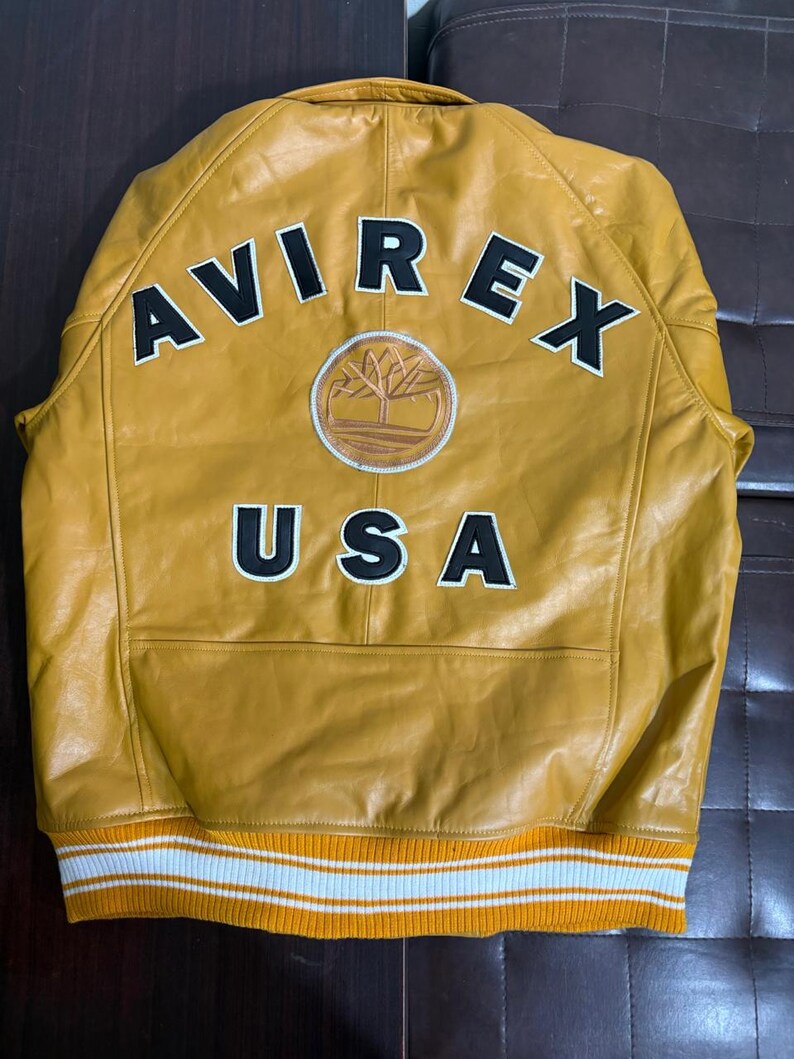 May include: Mustard yellow leather jacket with the text "AVIREX USA" in black and white. Features a circular patch with a tree design. The jacket has a ribbed knit hem with white and orange stripes.