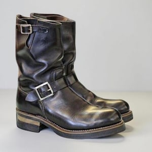 May include: A pair of black leather engineer boots featuring a mid-calf height, a rounded toe, and a stacked heel. Each boot includes a silver-toned metal buckle at the ankle strap and a smaller matching buckle at the top outer shaft.