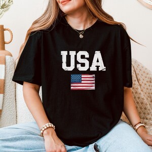 Pode incluir: T-shirt preto com a palavra "USA" em letras brancas desgastadas e uma pequena bandeira americana. A t-shirt tem gola redonda.