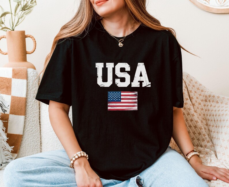 Pode incluir: T-shirt preto com a palavra "USA" em letras brancas desgastadas e uma pequena bandeira americana. A t-shirt tem gola redonda.