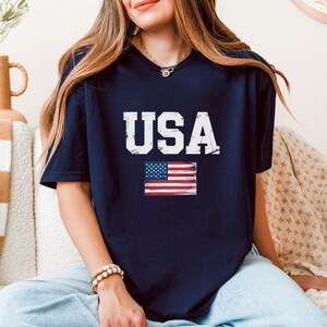 Pode incluir: Camiseta azul marinho com a palavra "USA" em letras brancas e uma bandeira americana desgastada. A camiseta tem decote em V e &eacute; feita de um material macio.