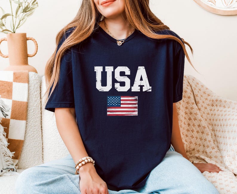 Pode incluir: Camiseta azul marinho com a palavra "USA" em letras brancas e uma bandeira americana desgastada. A camiseta tem decote em V e &eacute; feita de um material macio.