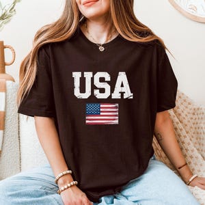 Pode incluir: Uma camiseta marrom escuro com a palavra "USA" em letras brancas desgastadas e uma pequena bandeira americana. A camiseta casual &eacute; adequada para o uso di&aacute;rio.