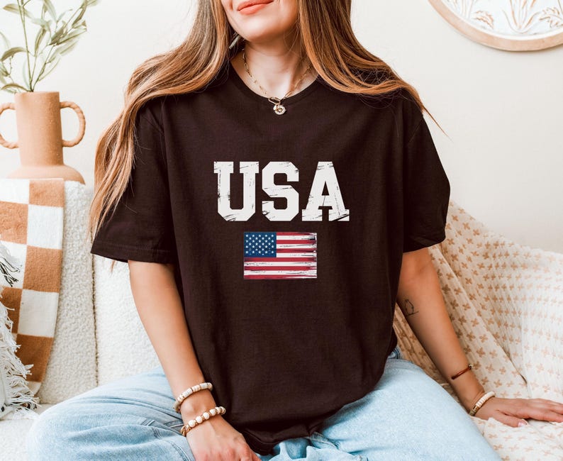 Pode incluir: Uma camiseta marrom escuro com a palavra "USA" em letras brancas desgastadas e uma pequena bandeira americana. A camiseta casual &eacute; adequada para o uso di&aacute;rio.
