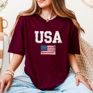 Pode incluir: Camiseta bord&ocirc; com a palavra "USA" em letras brancas desgastadas e uma pequena bandeira americana. A camiseta tem mangas curtas e gola redonda. O estilo geral &eacute; casual e patri&oacute;tico.