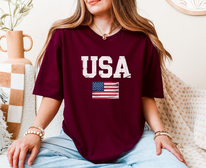 Pode incluir: Camiseta bord&ocirc; com a palavra "USA" em letras brancas desgastadas e uma pequena bandeira americana. A camiseta tem mangas curtas e gola redonda. O estilo geral &eacute; casual e patri&oacute;tico.