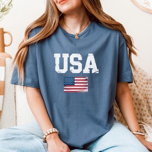 Pode incluir: Uma camiseta azul com a palavra "USA" em letras brancas e uma bandeira americana desgastada. A camiseta &eacute; feita de um material macio.