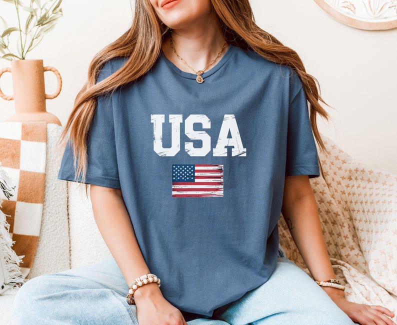 Pode incluir: Uma camiseta azul com a palavra "USA" em letras brancas e uma bandeira americana desgastada. A camiseta &eacute; feita de um material macio.