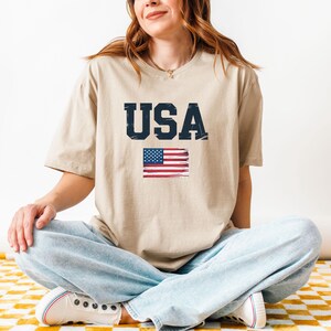 Pode incluir: Camiseta bege com a palavra "USA" em fonte azul marinho desgastada e uma bandeira americana pintada. A camiseta &eacute; de estilo casual.