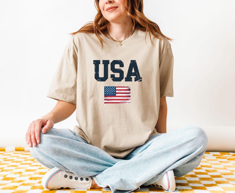 Pode incluir: Camiseta bege com a palavra "USA" em fonte azul marinho desgastada e uma bandeira americana pintada. A camiseta &eacute; de estilo casual.