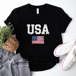 Pode incluir: T-shirt preto com a palavra "USA" em branco, com letras desgastadas e uma pequena bandeira americana. A imagem inclui jeans cinzentos, uma cesta de vime com vegeta&ccedil;&atilde;o e t&ecirc;nis brancos.