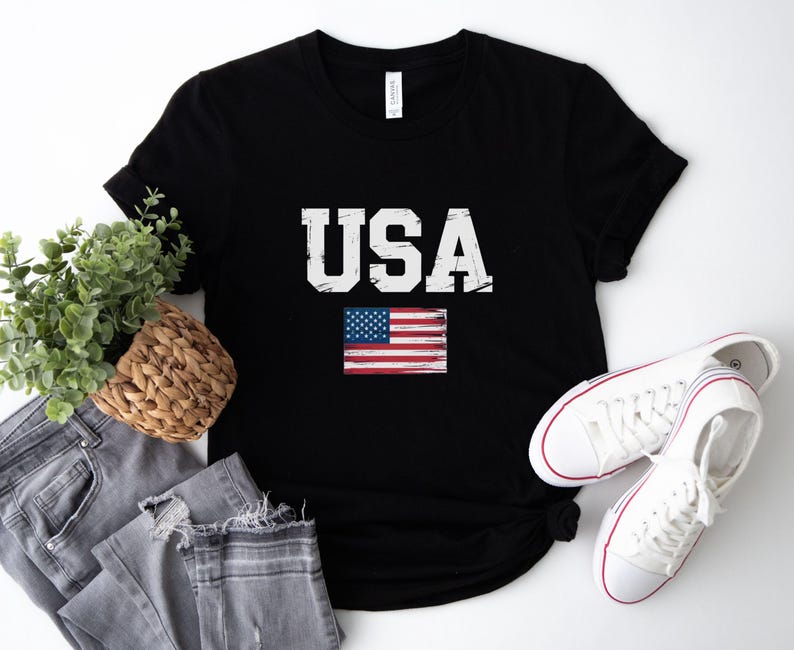 Pode incluir: T-shirt preto com a palavra "USA" em branco, com letras desgastadas e uma pequena bandeira americana. A imagem inclui jeans cinzentos, uma cesta de vime com vegeta&ccedil;&atilde;o e t&ecirc;nis brancos.