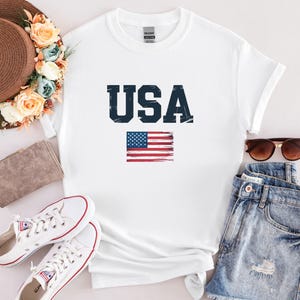 Pode incluir: Camiseta branca com a palavra "USA" em azul marinho e um design desgastado da bandeira americana. A camiseta &eacute; combinada com shorts jeans, &oacute;culos de sol e t&ecirc;nis brancos.