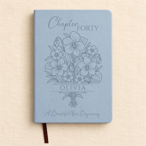 May include: A light blue hardback journal featuring a floral bouquet illustration in the centre. The text reads Chapter FORTY, OLIVIA, and A Beautiful New Beginning. A dark brown ribbon bookmark extends from the bottom of the rectangular notebook.