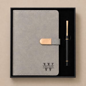 May include: A grey notebook with a smooth finish and a gold-toned metal clasp closure. The cover features the name WILLIAM engraved above a stylised W monogram. A black pen with gold-toned accents is positioned in a black tray next to the notebook.