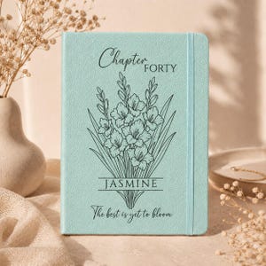 May include: A light mint green notebook with a textured cover featuring a black line art illustration of a bouquet of gladiolus flowers. The text reads Chapter FORTY, JASMINE, and The best is yet to bloom. It includes a vertical elastic band.