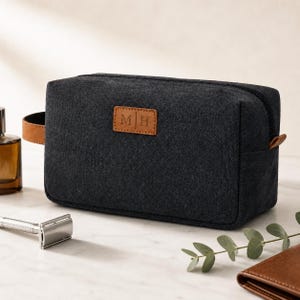 May include: A rectangular charcoal grey toiletry bag made of canvas fabric with a black zip closure. The front features a tan leather patch embossed with the letters M H. A tan leather side handle is attached to the left side of the bag.