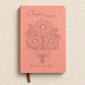 May include: A coral-coloured notebook featuring a line-art illustration of a daisy bouquet. The cover text reads Chapter THIRTY, OLIVIA, and The best is yet to bloom. A small brown ribbon bookmark extends from the bottom edge of the rectangular journal.