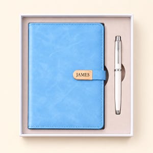 Op de afbeelding: Een lichtblauw notitieboek met een magnetische sluiting voorzien van een goudkleurig metalen plaatje met de inscriptie JAMES, gecombineerd met een witte en zilveren balpen, gepresenteerd in een witte rechthoekige geschenkdoos.