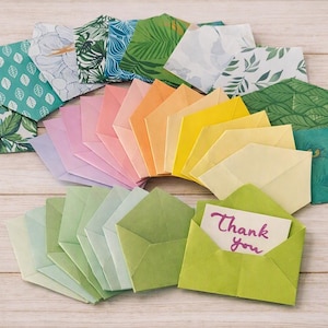 May include: A collection of small paper envelopes in a spectrum of colors including green, yellow, orange, pink, and purple, featuring various botanical and peacock feather patterns. One light green envelope contains a card with the text Thank you.
