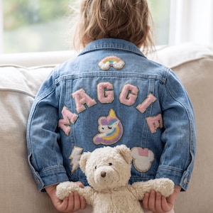 Puede incluir: Una chaqueta de mezclilla azul con el nombre MAGGIE en letras de tela rosa en la espalda. La chaqueta está decorada con parches bordados que incluyen un arcoíris, una cabeza de unicornio, un rayo y una forma de orejas de ratón, junto a un oso de peluche.