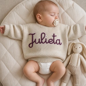 Puede incluir: Un suéter de punto de manga larga color crema con el nombre Julieta bordado en hilo morado oscuro en el pecho, acentuado con un pequeño detalle de flor blanca. El suéter lo lleva puesto un bebé dormido con un chupete y un juguete de peluche.