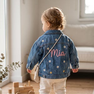 May include: A blue denim jacket featuring a collar, long sleeves, and buttoned cuffs. The back of the jacket is decorated with embroidered white and yellow daisy flowers and the name Mia stitched in pink script lettering across the centre.
