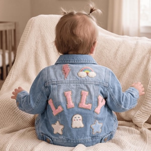 May include: A light blue denim jacket featuring the name LILY in large pink fabric letters on the back. The jacket is decorated with embroidered patches including a pink lightning bolt, a rainbow, a white ghost, and two stars in white and blue.