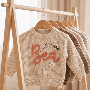 Könnte beinhalten: Ein cremefarbener, gesprenkelter Strickpullover mit dem in rosa Garn gestickten Namen Bea, ergänzt durch eine kleine Biene und eine weiße Pompon-Blumenapplikation. Der Pullover hängt an einem Holzbügel auf einem Kleiderständer aus hellem Holz.