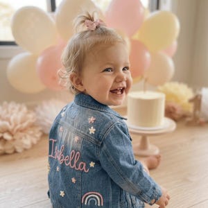 May include: A blue denim jacket featuring embroidered details on the back, including the name Isabella in pink script, small white and pink flowers, a yellow sun, and a pastel-coloured rainbow. The jacket has a classic collar and button cuffs.