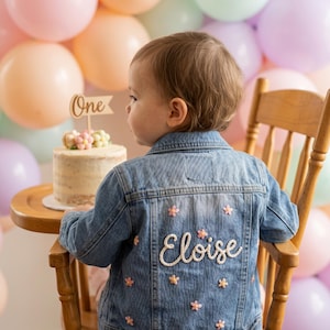 May include: A blue denim jacket featuring white embroidered script text that reads Eloise on the back, accented with small pink and yellow floral embroidery throughout the fabric. The jacket is worn by a child sitting in a wooden chair.