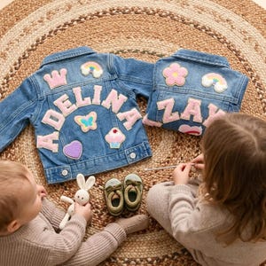 May include: Two denim jackets with patches and names, "ADELINA" and "ZAY", are displayed on a woven rug. The jackets have pink patches of a crown, rainbow, flower, butterfly, heart, and cupcake. A child holds a crochet bunny and another child is working on a craft.