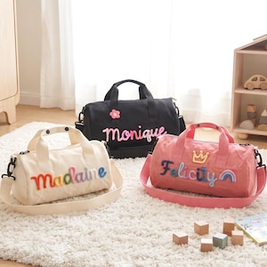 May include: Three cylindrical duffel bags in black, cream, and pink. The black bag features the name Monique and a pink flower. The cream bag displays the name Madaine in rainbow colors. The pink bag shows the name Felicity with a crown and rainbow.
