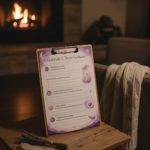 Puede incluir: Un portapapeles de madera con una hoja impresa titulada Gratitude & Divine Guidance. El papel presenta gráficos celestiales en morado y dorado, fases lunares y secciones con líneas para registrar gratitud y guía divina.