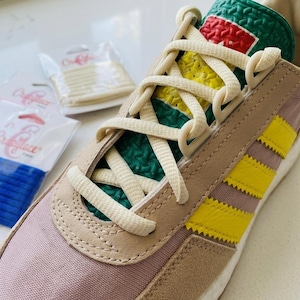 Peut inclure: Une basket basse avec une tige en toile et daim mauve ornée de trois bandes latérales jaune vif. La languette présente un motif texturé vert, jaune et rouge. Des lacets crème sont passés dans les œillets, avec deux petits paquets en plastique de lacets de rechange visibles en arrière-plan.