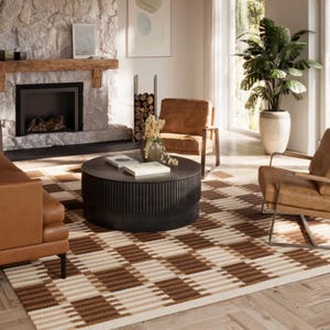 May include: A large, round, black coffee table with a vertically ribbed base, positioned on a brown and cream checkered area rug. The table surface holds two white books and a small vase with dried flowers, centered in a living room.
