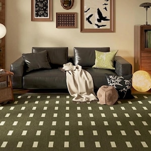 May include: A rectangular area rug featuring a dark olive green base with a repeating pattern of small, light-colored vertical rectangles arranged in a grid. The rug is positioned in front of a dark leather sofa with various throw pillows and a blanket.
