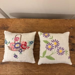 May include: Two small, square, off-white throw cushions with embroidered floral designs. One cushion features a basket of colourful flowers, the other has purple daisies with yellow centres and green leaves. The cushions are on a wooden surface.