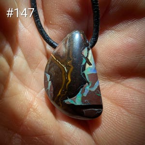 Peut inclure: Un pendentif en opale boulder polie avec une matrice en pierre de fer brun foncé et des éclats vibrants de couleur irisée bleue et verte, suspendu à un cordon noir. La pierre présente une forme irrégulière et le texte #147 dans le coin.
