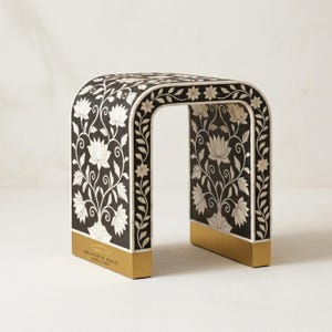 May include: A decorative black and white floral patterned side table with a gold base. The table features a curved design with a rectangular top and a cutout center. The floral pattern includes lotus flowers and vines.