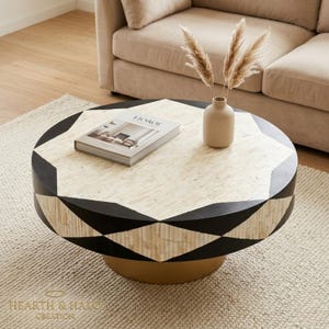 Puede incluir: Una mesa de centro redonda con un patrón geométrico de incrustaciones de hueso color crema y segmentos de resina negra. La mesa tiene una base dorada y sostiene un jarrón de cerámica beige con hierba de la pampa seca y un libro titulado HOROE.