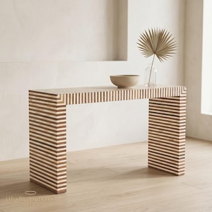 Puede incluir: Una consola rectangular con un patrón de rayas en tonos marrón y crema. La mesa tiene un diseño minimalista con una parte superior plana y dos patas de soporte. Un cuenco beige y un jarrón transparente con una hoja de palma seca encima.