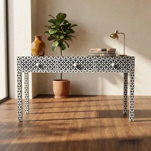 May include: A black and white patterned console table with three drawers and white knobs. The table has a geometric design. A small gold lamp, books, and a vase are on top of the table. The table is 120 cm long.