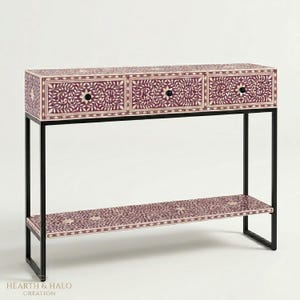 May include: A console table with a black metal frame and a patterned top and shelf. The top has three drawers with black knobs and a floral design in purple and cream. The shelf below mirrors the top's design, offering additional storage.