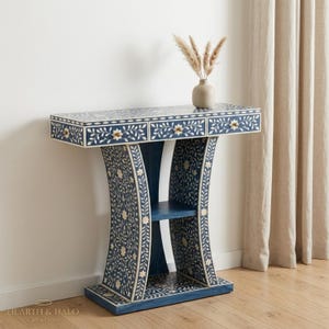 May include: A navy blue console table featuring intricate white floral and vine inlay patterns across its surface. The piece includes three drawers with small flower-shaped knobs and a curved base design with an open shelf. A beige vase sits on top.