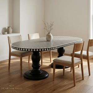Puede incluir: Una mesa de comedor ovalada con una superficie de rayas blancas y negras y un borde con patrón de diamantes, sostenida por dos bases de pedestal negras. Cuatro sillas de madera clara con asientos tapizados en beige rodean la mesa.