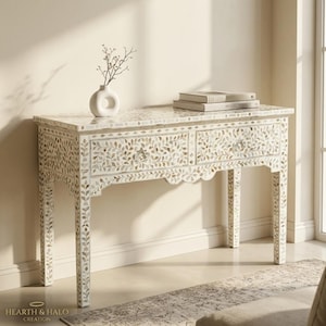 May include: A rectangular console table featuring an intricate floral inlay pattern made of iridescent shell pieces. The table has two drawers with small knobs, four straight legs, and a white ceramic vase with thin branches on top.
