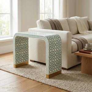 May include: A waterfall console table featuring a repeating geometric pattern in sage green and white. The table has a curved silhouette with solid gold-toned metal bases at the bottom of each side, providing a stable foundation for the piece.