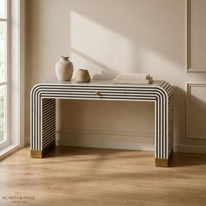 May include: A rectangular console table featuring a black and white striped pattern with rounded corners, a single central drawer with a small gold knob, and gold-toned metal base accents. A beige ceramic vase and a woven bowl sit on the top surface.