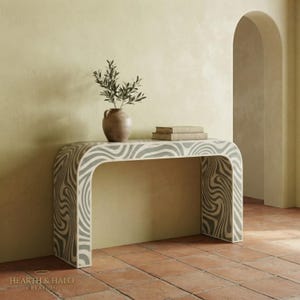 May include: A console table with a curved arch shape and a gray and white wavy marble-like pattern. On top sits a brown ceramic vase containing a green leafy plant and two stacked books. The text HEARTH & HALO CREATION is visible at the bottom.